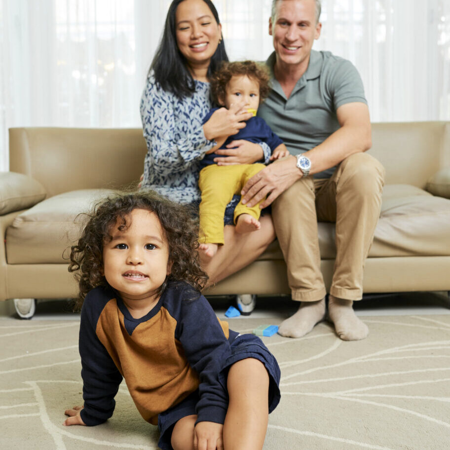 Happy Kid and His Family Cute positive multi-ethnic kid sitting on he floor, his parents and little brother sitting on sofa in background